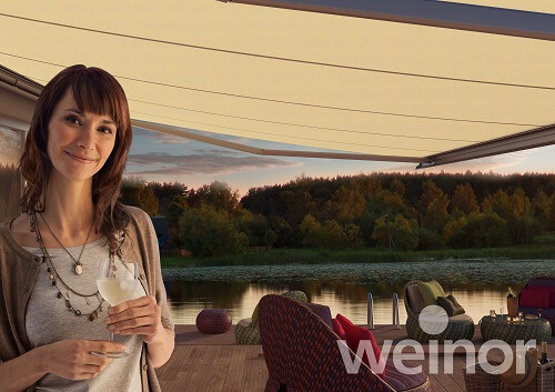 Frau genießt Getränk auf Terrasse mit stilvoller Markise am See bei Sonnenuntergang.